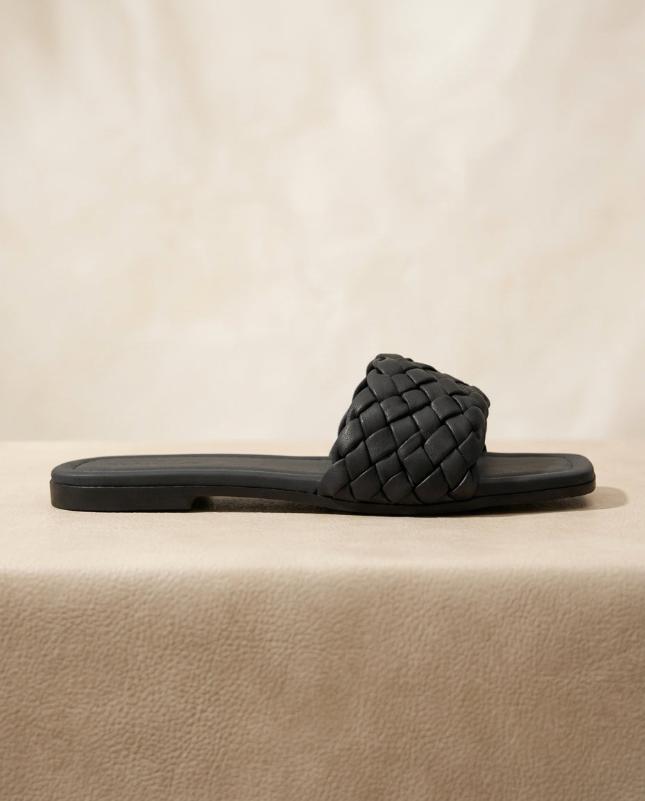 "Minimalist studio photography of a single UHR Collection women's black braided flat slide sandal, shot from a clean side profile angle on a textured beige matte surface."