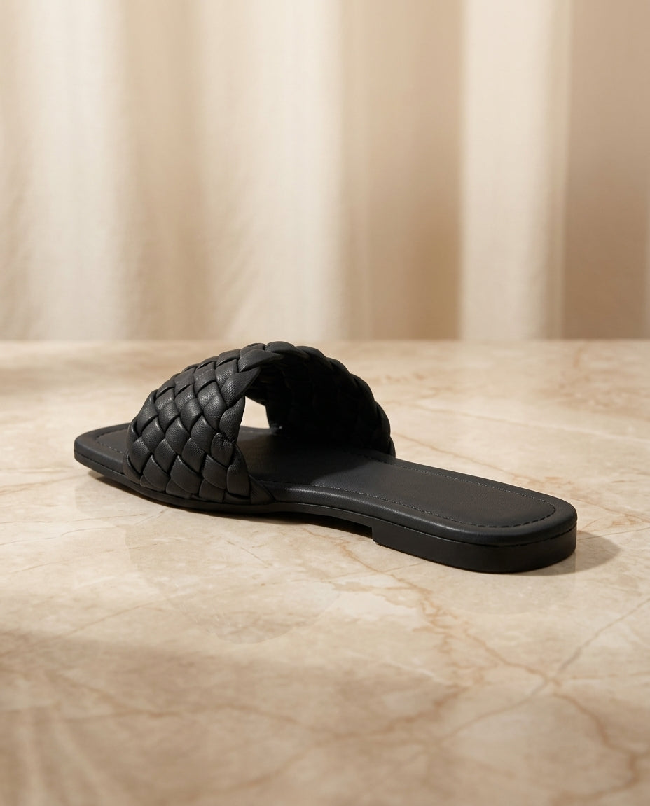 "Minimalist studio photography of a single UHR Collection women's black braided flat slide sandal, shot from a clean side profile angle on a textured beige matte surface."